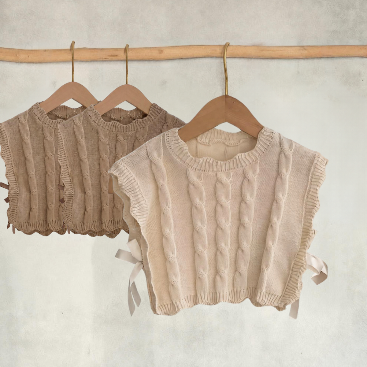Cream girls’ cable-knit vest with side ribbon ties in focus, with two light brown vests displayed behind on wooden hangers