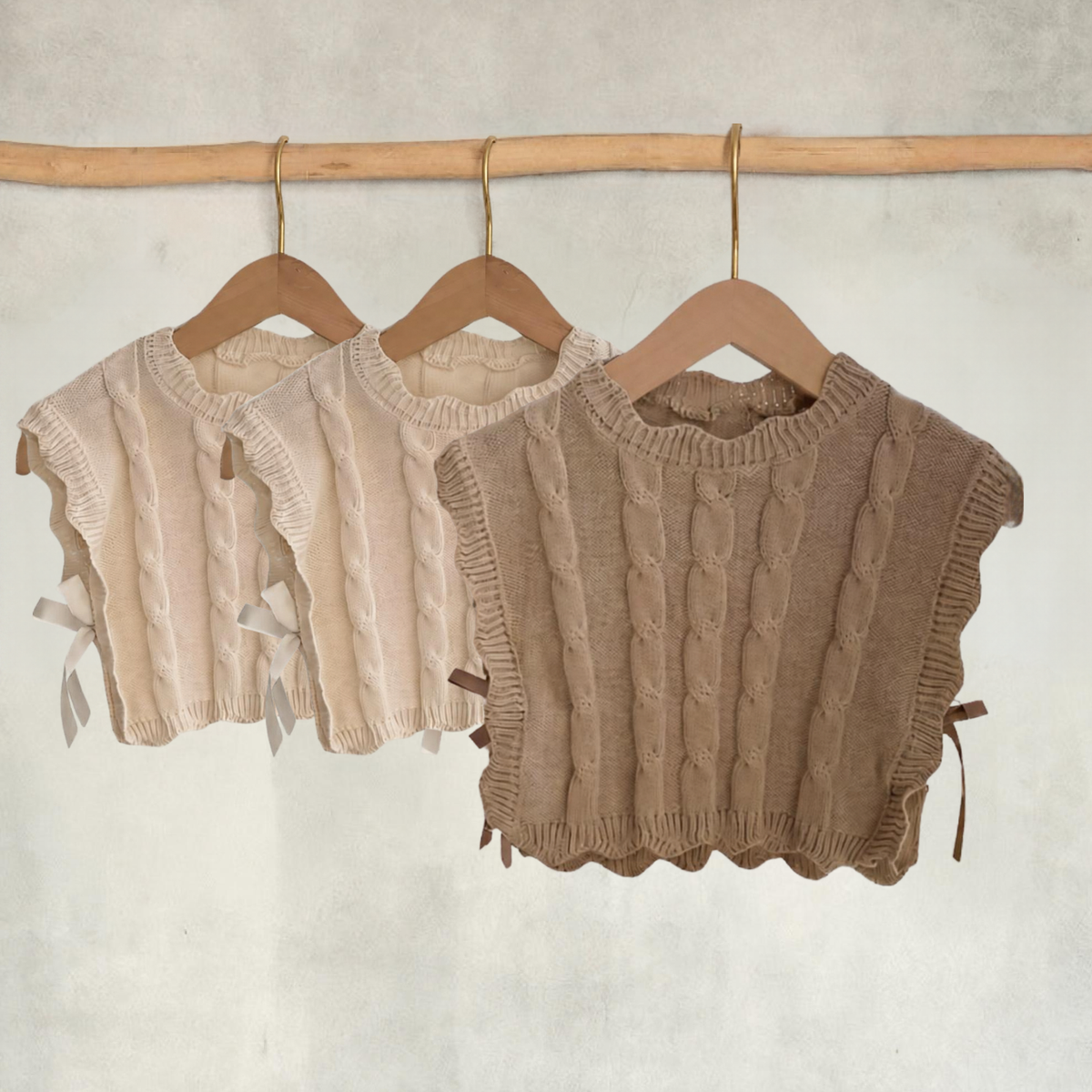 Light brown girls’ cable-knit vest with side ribbon ties in focus, with two cream vests displayed behind on wooden hangers