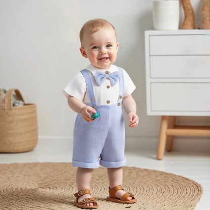 Baby boy wearing a powder blue suspender romper set standing indoors.