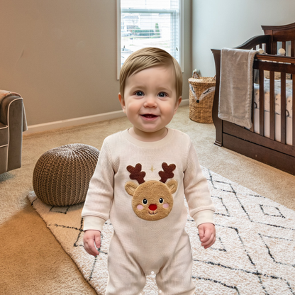 Baby standing in a neutral nursery wearing a cream waffle-textured reindeer romper featuring brown antlers, a red nose and a raised plush appliqué