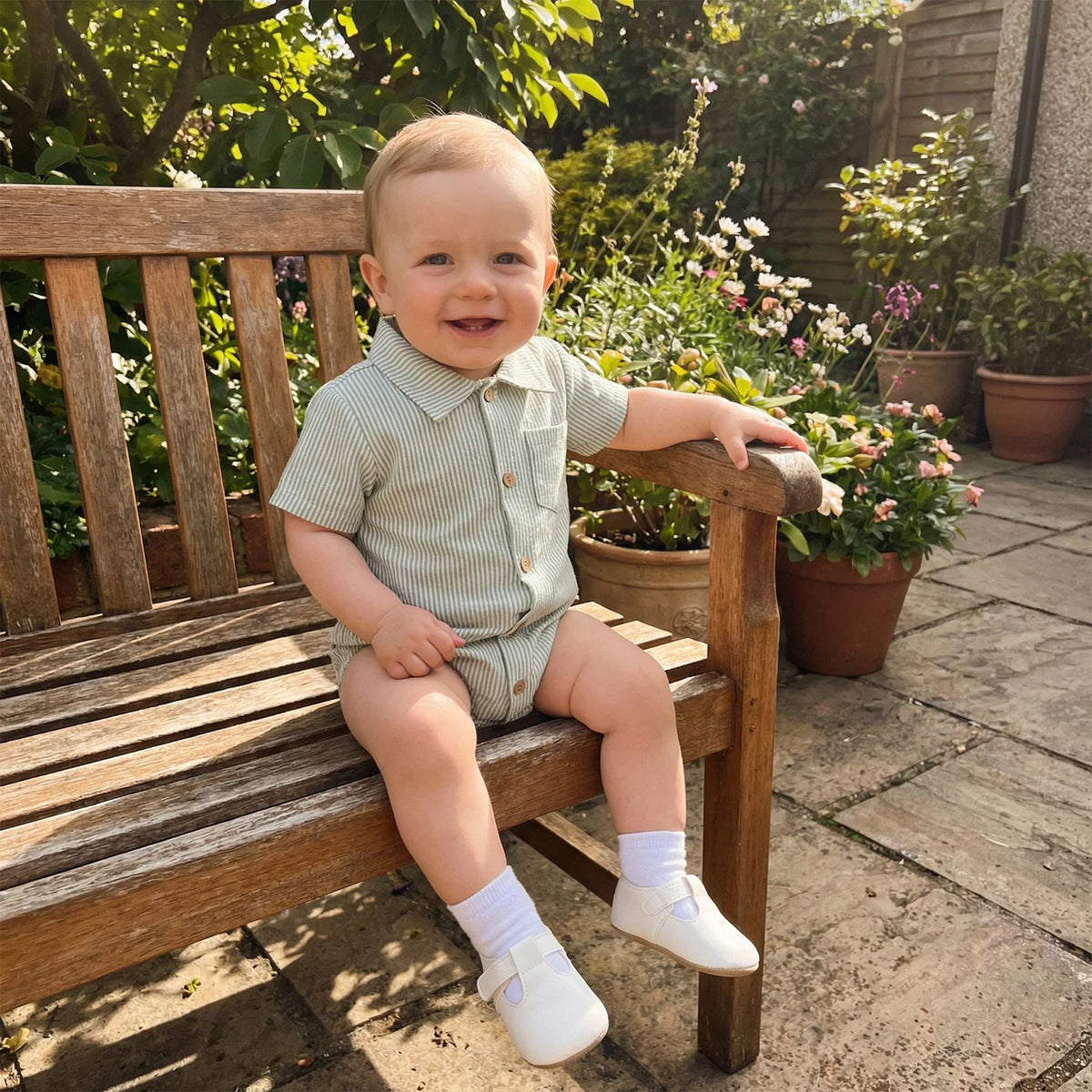 Happy baby wearing a sage green striped cotton romper sitting on a wooden bench outdoors.