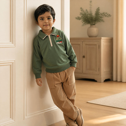 Toddler boy wearing the Little Evergreen Christmas Tree Quarter-Zip Pullover in green, leaning against an ivory paneled wall in a cozy neutral room