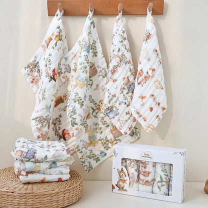 “Set of luxury muslin baby burp cloths in whimsical woodland animal prints, displayed hanging on wooden hooks with gift box and folded cloths – soft, breathable cotton gauze for newborn care and gifting.”