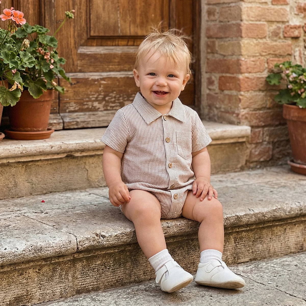 Smiling baby wearing a beige striped cotton romper sitting on stone steps.