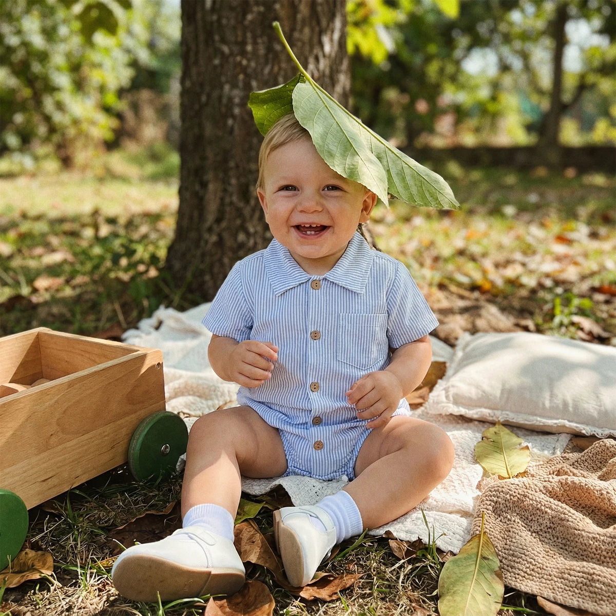 Smiling baby wearing a sky blue striped cotton romper sitting outdoors under a tree.