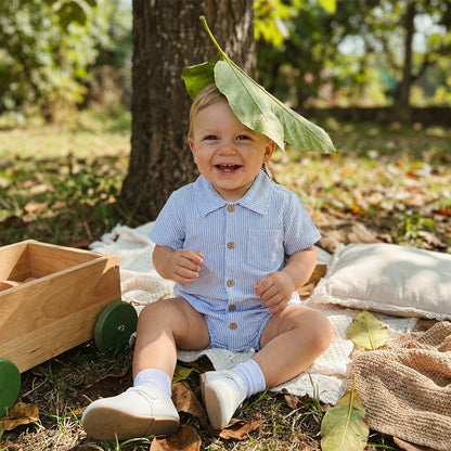 Smiling baby wearing a sky blue striped cotton romper sitting outdoors under a tree.