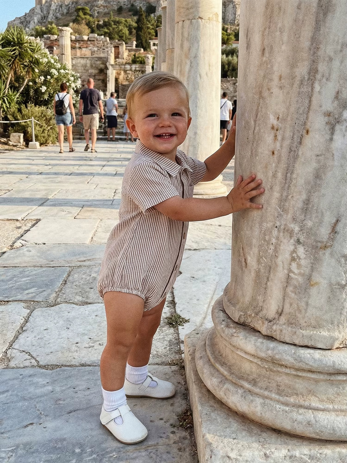 Baby wearing a beige striped cotton romper standing beside a stone column.